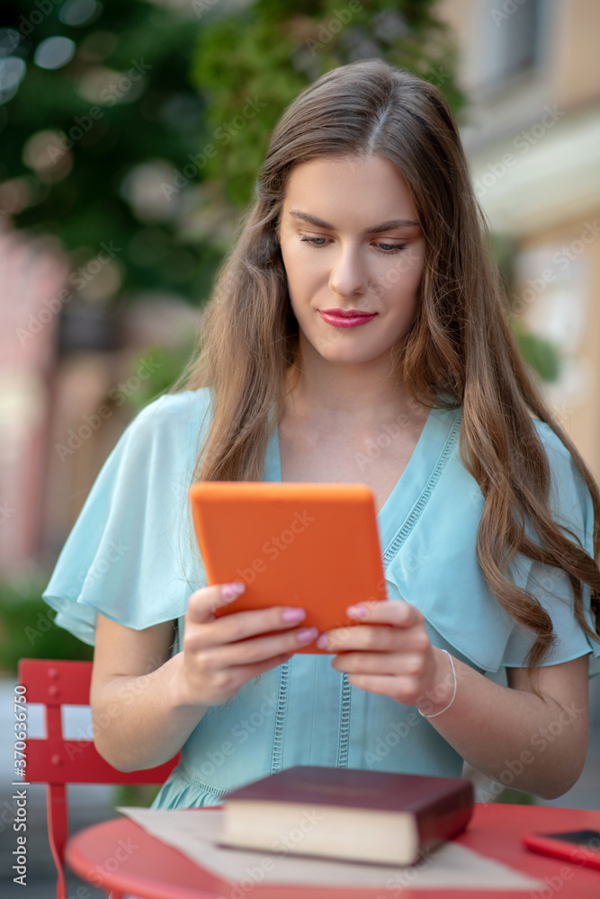 Obraz premium Female in blue dress sitting in cafe, holding orange tablet, looking at screen