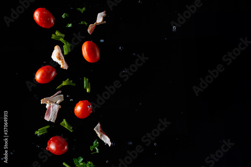 Falling pieces of fresh bacon, cherry tomatoes, parsley and onions on a black background, freeze in motion, for design, cooking
