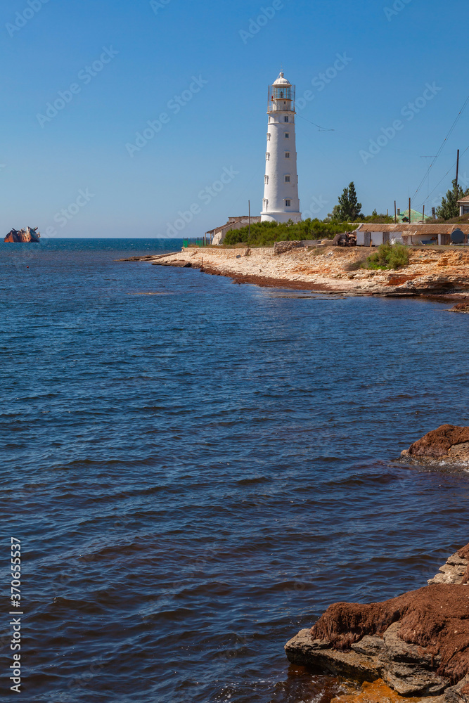 The white lighthouse stands on the seashore. Not far from it is a grounded ship.
