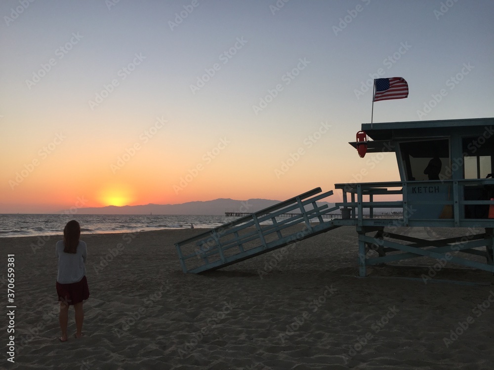 Female tourist overlooking landscape view of empty coastal beach at sunset near lifeguard tower