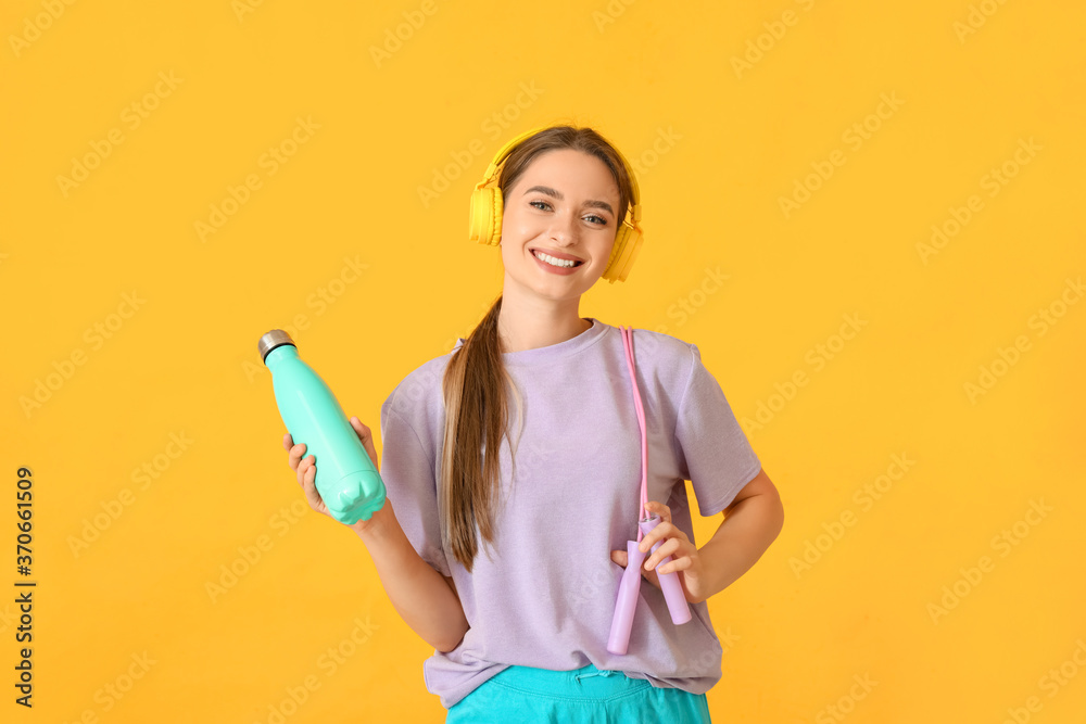 Sporty young woman with headphones and water on color background