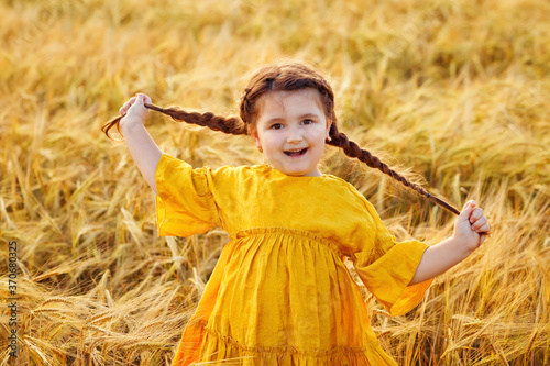 Portrait of a beautiful girl in a yellow dress and two pigtails on her head on a Golden wheat field at sunset. The girl is having fun in the fall.