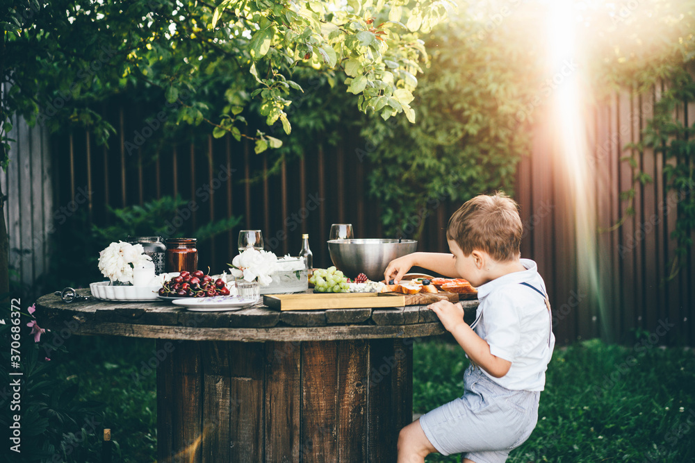 funny little boy in white shirt takes fresh blueberry from toast at table with festive setting in summerhouse green backyard. - obrazy, fototapety, plakaty 