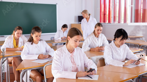 Medical students using mobile phones and writing in notepads during lesson