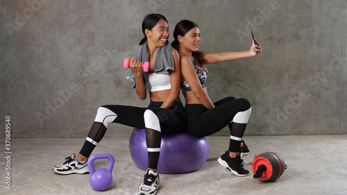 Two beautiful young Asian women posing with fitness ball and dumbbell while doing selfie photo at the gym. Happy female friends spending their free time at the sport centre