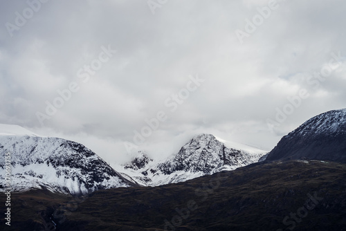 Great black mountains with white snow on tops and glaciers. Dramatic landscape with snowy mountains under cloudy gray sky. Atmospheric alpine scenery with snow on rocky mountains in overcast weather.