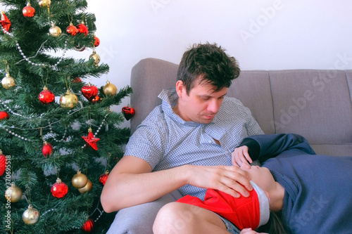 Couple man and woman sleeping and waiking together on couch near Christmas tree at home. New Year night on winter holidays. Morning hangover after party. Family life. Resting hugging each other. photo