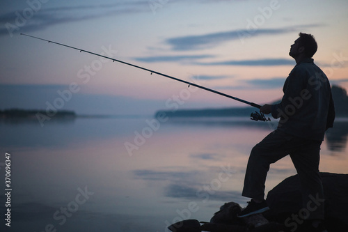 Fisher man fishing with spinning rod on a river bank at misty foggy dusk