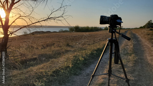 DSLR photographer equipment while capture picture of sunset over lake. Professional photo camera on tripod stands at road near bare tree and looks to sunset sun over water. Countryside landscape
