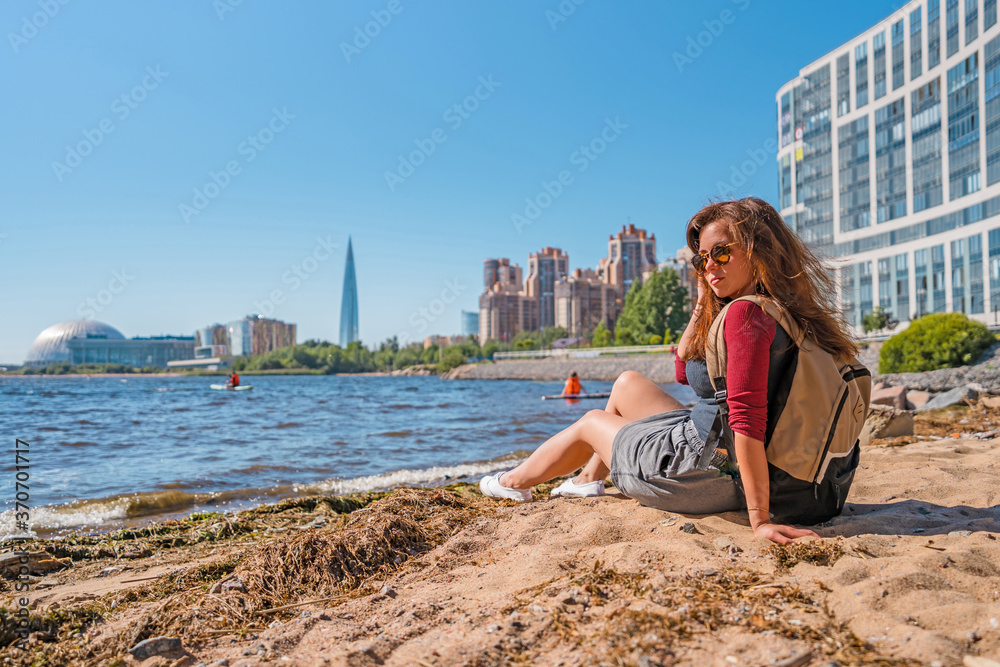 A young woman with a backpack made a stop on the beach of Saint Petersburg on the Gulf of Finland with a view of business centers and the Lakhta Center skyscraper