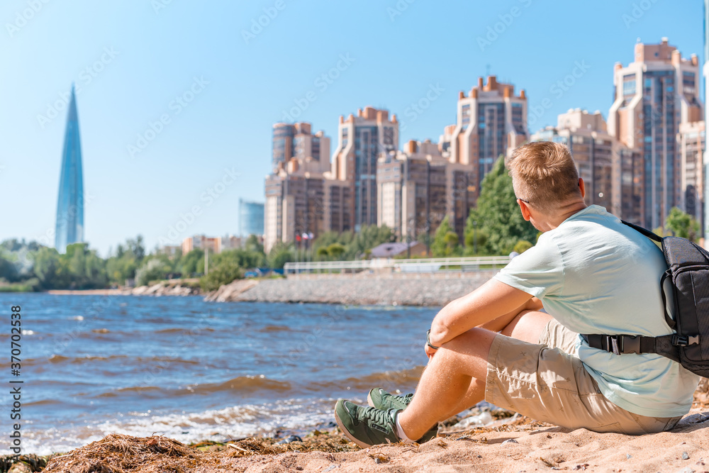 A young man with a backpack made a stop on the beach of Saint Petersburg on the Gulf of Finland with a view of business centers and the Lakhta Center skyscraper