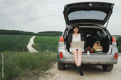 Young Stylish Woman Sits on the Trunk of Her SUV Car and Working on a Laptop in the Green Field