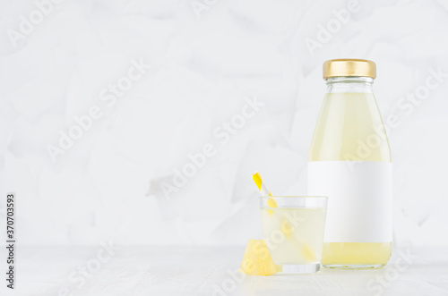 Summer yellow pineapple juice in glass bottle mock up with blank label, straw, wine glass, fruit slice on white wood table in light interior, template for packaging, advertising.