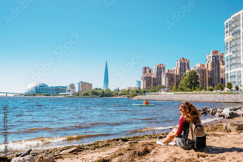A girl sits on the beach by the sea of the Gulf of Finland in St. Petersburg with a view of the Lakhta Center skyscraper