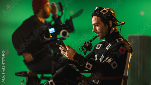 Handsome Smiling Actor Wearing Motion Capture Suit and Head Rig having Lunch Break, Sitting on Chair, Uses Smartphone. Studio High Budget Movie. On Film Studio Period Costume Drama Film Set photo