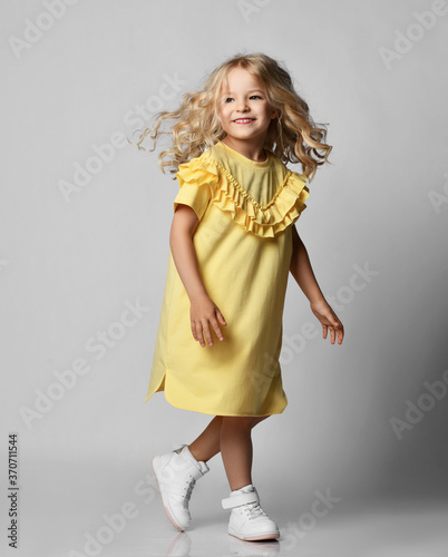 Canvas Print Frolic smiling blonde curly kid girl in yellow dress and sneakers walking moving over grey wall background