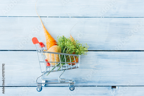 small metal grocery cart full of farm vegetables: onions, carrots, cucumber, dill on blue wooden background, top view, space for text