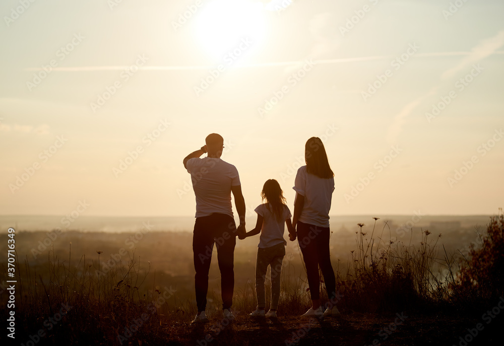 Silhouette of a family - father, mother and daughter standing on the hill with their backs to the camera and looking to the horizon on the sunset Silhouette of a family - father, mother and daughter standing on the hill with their backs to the camera and looking to the horizon on the sunset