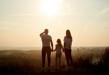 Silhouette of a family - father, mother and daughter standing on the hill with their backs to the camera and looking to the horizon on the sunset