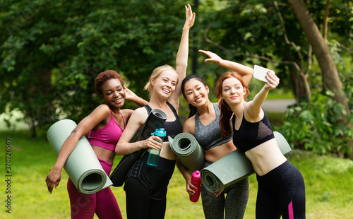 Cheerful multiracial girls in sports clothes taking selfie before their outdoor yoga practice