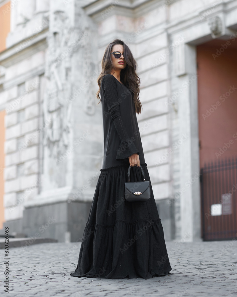 Young beautiful woman with brunette hair wearing long black dress and sunglasses, holding leather handbag and walking at city street on a summer day. Outdoor full length fashion portrait. Female model