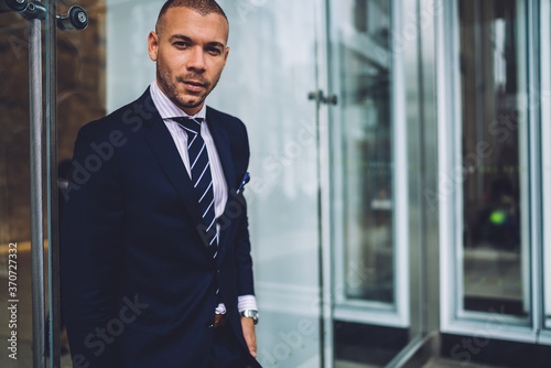 Portrait of self confident executive manager in stylish suit standing near copy space area for advertising, handsome serious businessman in formalwear looking at camera concentrated on career and job.