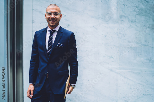 Prosperous male entrepreneur in elegant suit looking at camera satisfies with successful career standing on promotional background, portrait of smiling manager happy about setting up own business.