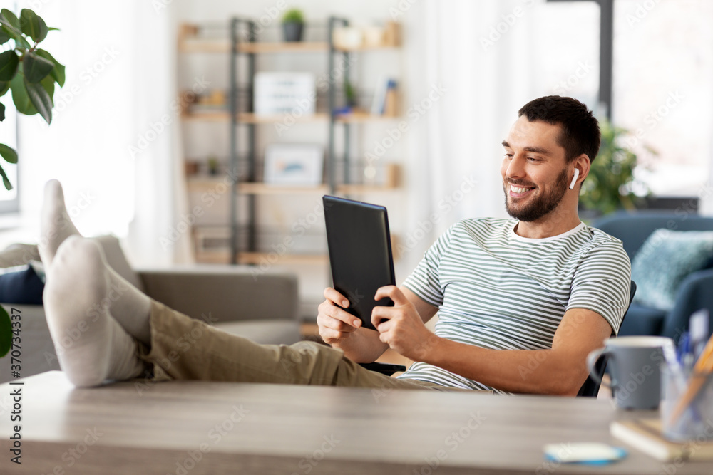 technology, remote job and lifestyle concept - happy smiling man with tablet pc computer and earphones resting feet on table at home office technology, remote job and lifestyle concept - happy smiling man with tablet pc computer and earphones resting feet on table at home office