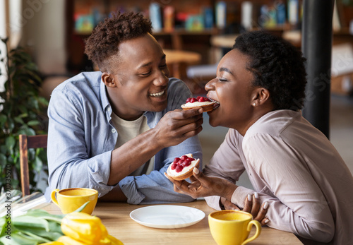 Millennial black man feeding tasty berry tartlet to his girlfriend at cozy cafe
