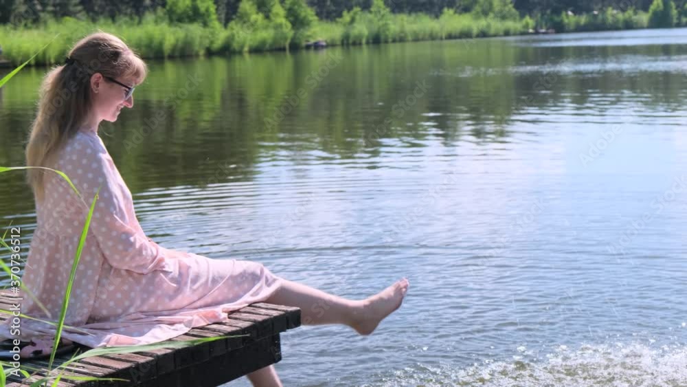 Woman on the lake. Young woman in pink dress sitting on a wooden platform on the lake and dip her feet in water, creating splashes, slow motion Woman on the lake. Young woman in pink dress sitting on a wooden platform on the lake and dip her feet in water, creating splashes, slow motion