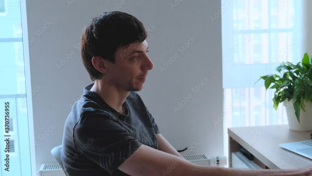 Side view of a young dark-haired man sitting at the table in front of her laptop telling something Side view of a young dark-haired man sitting at the table in front of her laptop telling something