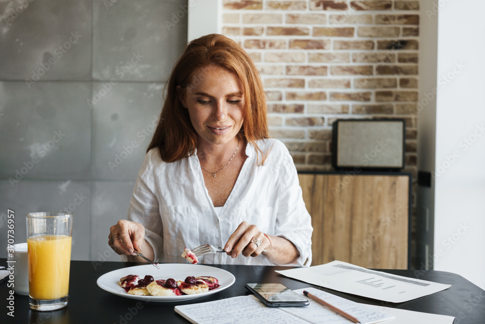 Image of pleased businesswoman using cellphone while having breakfast