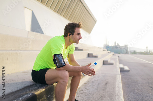 Young sports man listening to music in headphones while admiring sunrise and relaxing after an active run outdoors, handsome male jogger hold bottle of water and taking break after morning workout