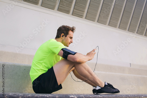 Side view portrait of caucasian male runner in headphones using smart phone for choose the music of playlist and continue his fit training,young sports man resting after his diary morning jog outdoors
