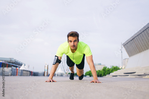 Handsome sports man doing warm up exercise before start his workout training outside, caucasian male jogger working out with hands leaning on asphalt city road while listening to music in headphones