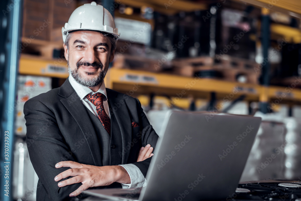 Businessman wearing a hardhat standing at goods warehouse and check for control loading from Cargo freight ship for import and export by report on laptop. commercial warehouse for trading concept - obrazy, fototapety, plakaty 