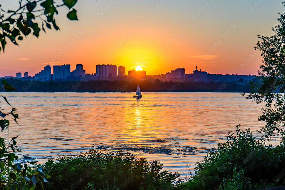 Fototapeta premium Sailing boat against the sunset behind modern buildings.