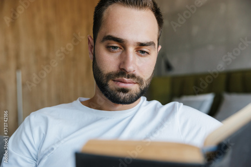 Image of young thinking man reading book while sitting on couch