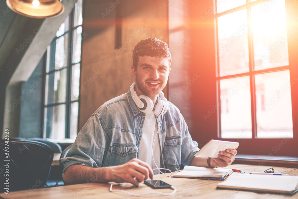 Portrait of cheerful hipster guy with touch pad in hand looking at camera while installing media application for listening music via electronic headphones, positive man using technology for chatting