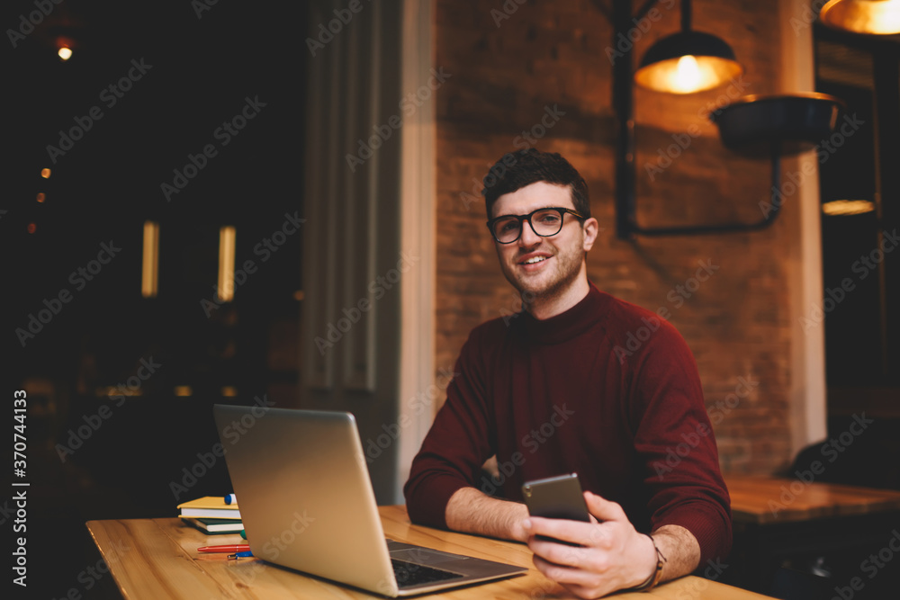Portrait of smiling hipster guy in spectacles looking at camera while having funny chatting conversation with friends using application on cellular while sitting in coworking space front laptop Portrait of smiling hipster guy in spectacles looking at camera while having funny chatting conversation with friends using application on cellular while sitting in coworking space front laptop