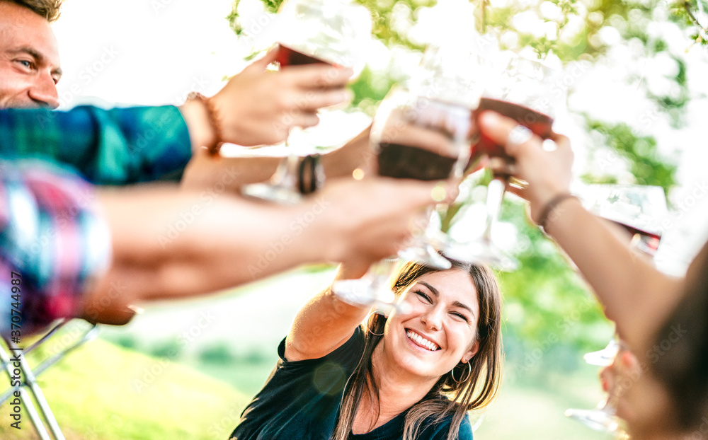 Naklejka premium Hands toast with red wine - People having fun cheering at pic nic winetasting - Young friends enjoying harvest time together at farmhouse vineyard countryside - Focus on woman with blurred glasses