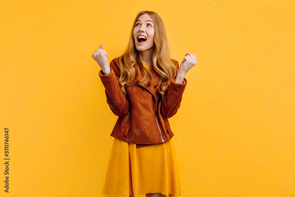 excited shocked screaming happy beautiful girl, in a bright yellow dress and autumn jacket, happily shows a winning gesture while standing on an yellow background. The concept of autumn excited shocked screaming happy beautiful girl, in a bright yellow dress and autumn jacket, happily shows a winning gesture while standing on an yellow background. The concept of autumn