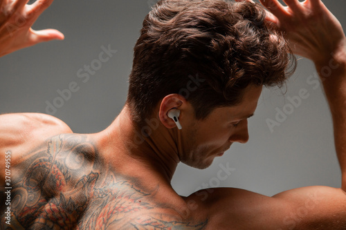 Cool young guy in a wireless earphone with stylish haircut listen to song without shirt. Music poster. Body with tattoo