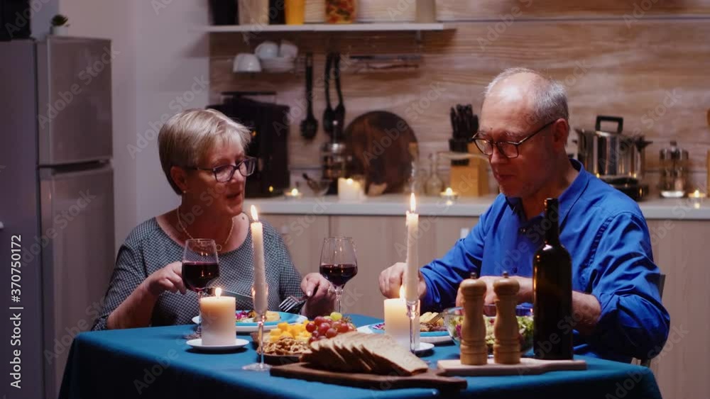Portrait of senior couple with red wine glasses sitting at the table in the cozy kitchen. Happy cheerful elderly couple dining together at home, enjoying the meal, celebrating their anniversary Portrait of senior couple with red wine glasses sitting at the table in the cozy kitchen. Happy cheerful elderly couple dining together at home, enjoying the meal, celebrating their anniversary