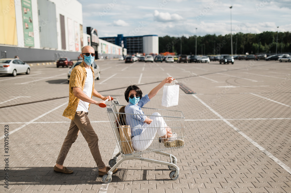 Obraz premium Shopping day. A carefree couple in love have fun during a shopping. The girl sits in a cart with paper bags and the guy carries the cart. They are wearing protective masks and sunglasses