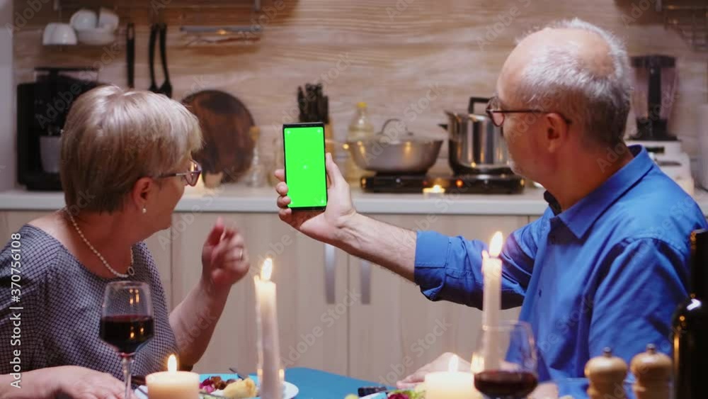 Old retired senior couple holding mockup phone at dinner. Aged people looking at green screen template chroma key isolated smart phone display using techology internet sitting at the table in kitchen.