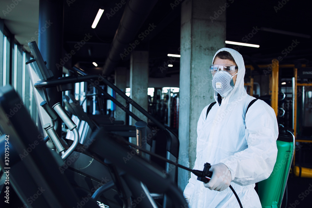 Gym disinfection of fitness equipment. Man in white protection suit disinfecting and spraying treadmill running tracks to stop spreading highly contagious coronavirus or COVID-19.