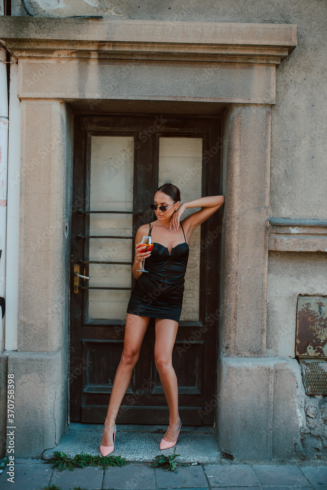 Full length of a beautiful attractive young caucasian woman in a black short dress with a cocktail in hand in front of the door