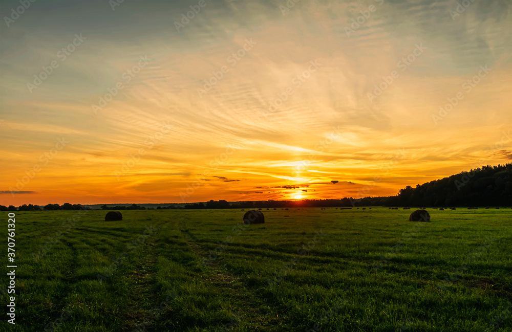 Scenic view at beautiful sunset in green shiny field with hay stacks, bright cloudy sky, country road and golden sun rays with glow, summer valley landscape