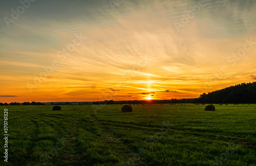 Scenic view at beautiful sunset in green shiny field with hay stacks, bright cloudy sky, country road and golden sun rays with glow, summer valley landscape © Yaroslav